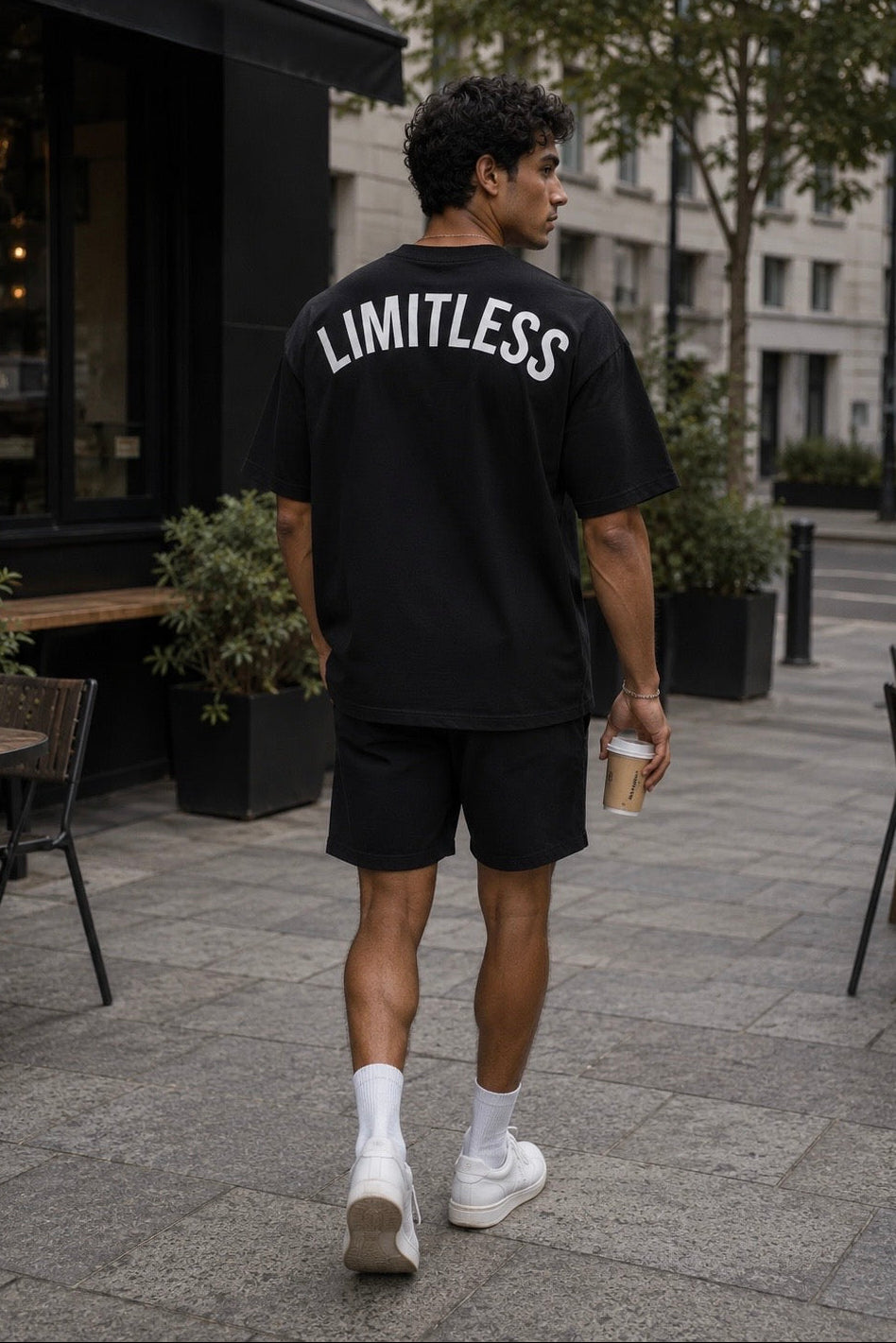 A man walking away on a city sidewalk wearing a black oversized T-shirt with 'LIMITLESS' printed in white across the back, black shorts, white socks, and white sneakers, while holding a coffee cup.
