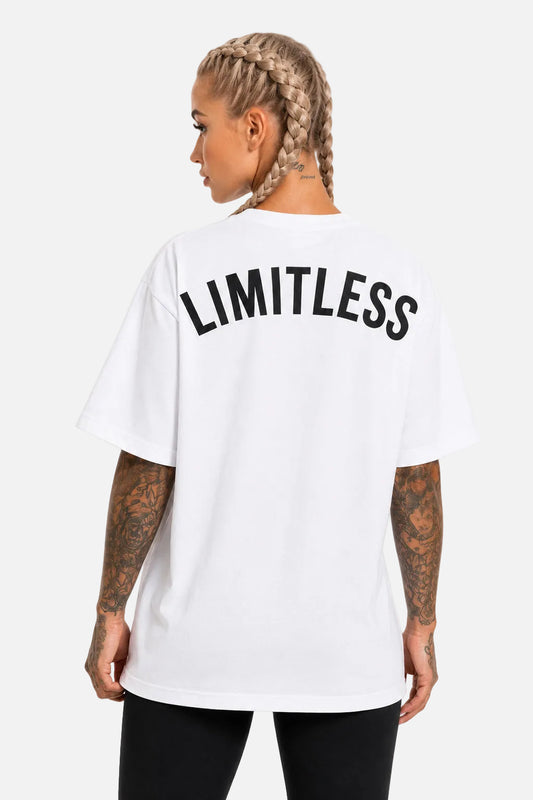 A woman shown from the back wearing a white oversized T-shirt with the word 'LIMITLESS' printed in large black letters across the upper back, paired with black pants against a plain white studio background.