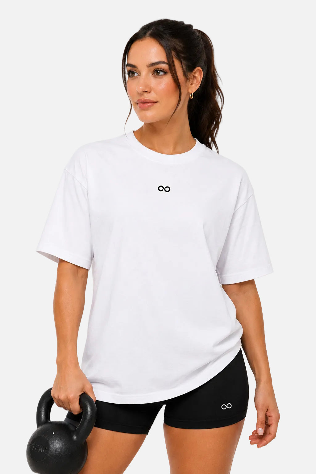A woman modeling a white short-sleeve crewneck t-shirt with a small black infinity symbol on the chest, paired with black athletic shorts and holding a black kettlebell against a solid white background.