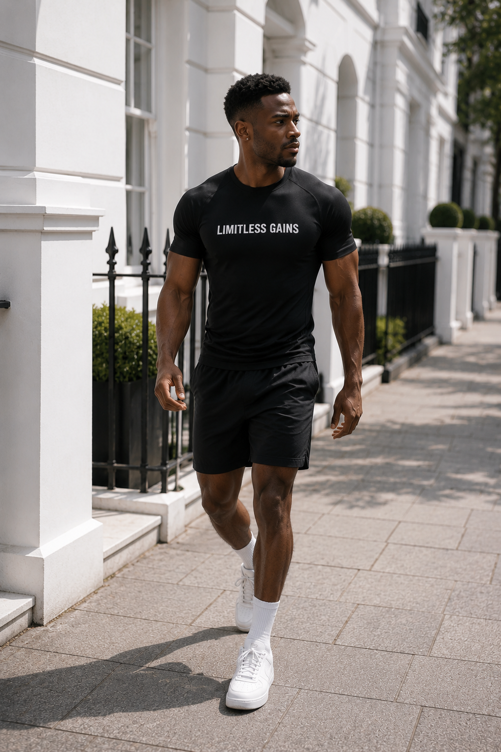 A man walking on a paved sidewalk in front of a white building, wearing a black t-shirt with 'LIMITLESS GAINS' printed in white across the chest, black athletic shorts, white socks, and white sneakers.