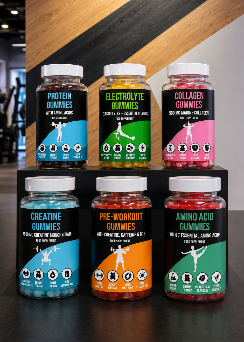 Six bottles of Anytime Fitness branded gummy supplements arranged on a black display stand in front of a wood-paneled wall with the Anytime Fitness logo. The bottles include Protein Gummies, Electrolyte Gummies, Collagen Gummies, Creatine Gummies, Pre-Workout Gummies, and Amino Acid Gummies, each with colorful labels and white caps.