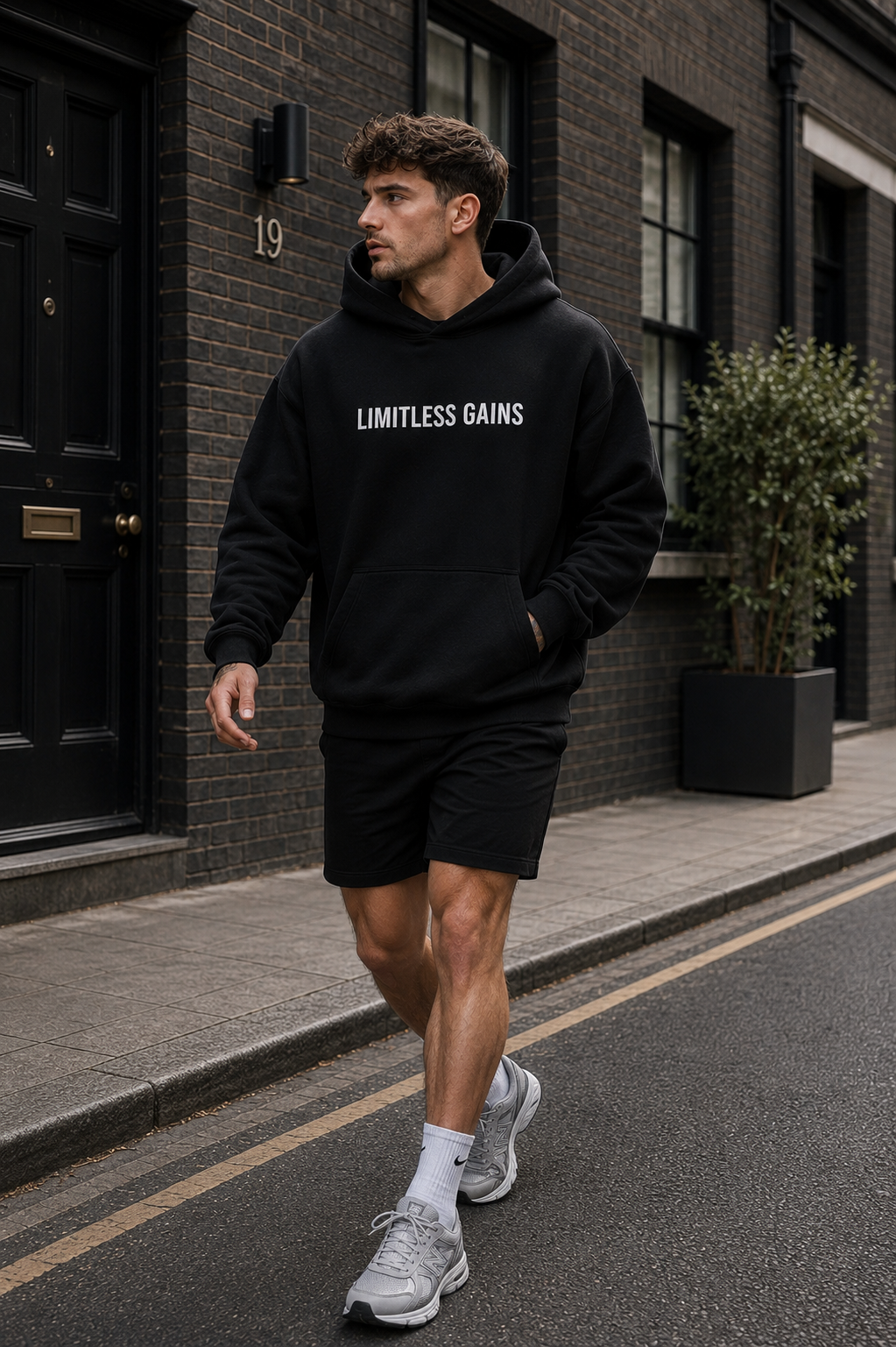A man walking on a city street wearing a black pullover hoodie with the words 'LIMITLESS GAINS' printed in white across the chest, paired with black shorts and gray sneakers.