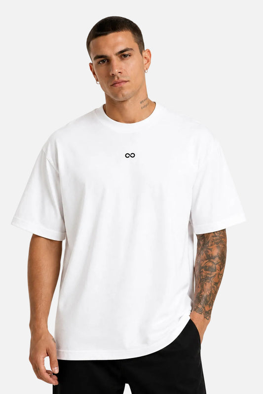 A man wearing a white short-sleeve crewneck t-shirt with a small black infinity symbol logo on the center chest, paired with black pants, against a plain white background.