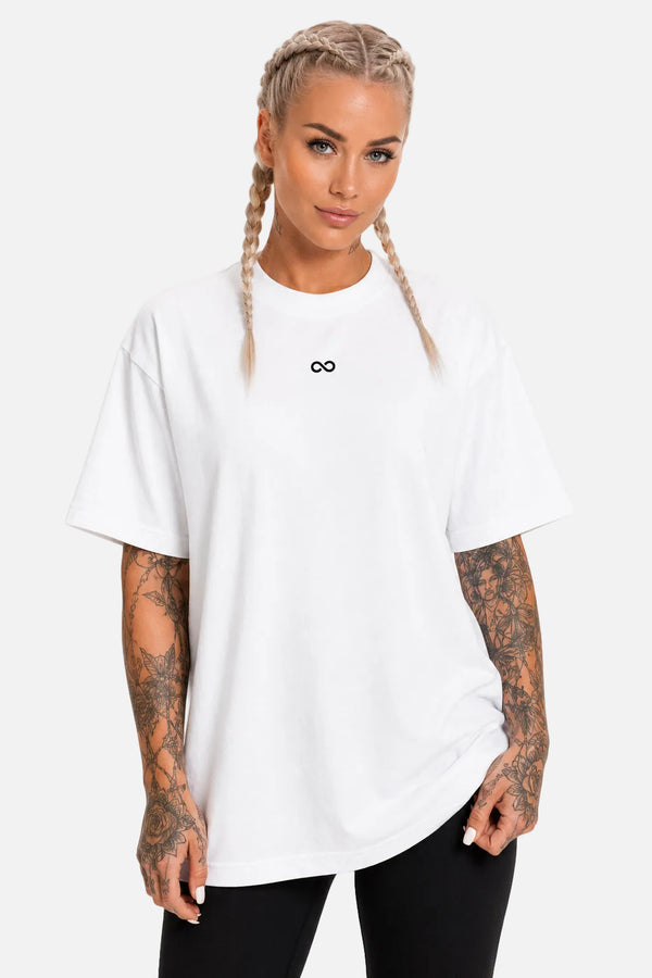 A woman with blonde braided hair wearing an oversized white short-sleeve t-shirt with a small black infinity symbol on the chest, paired with black pants, against a plain white background.